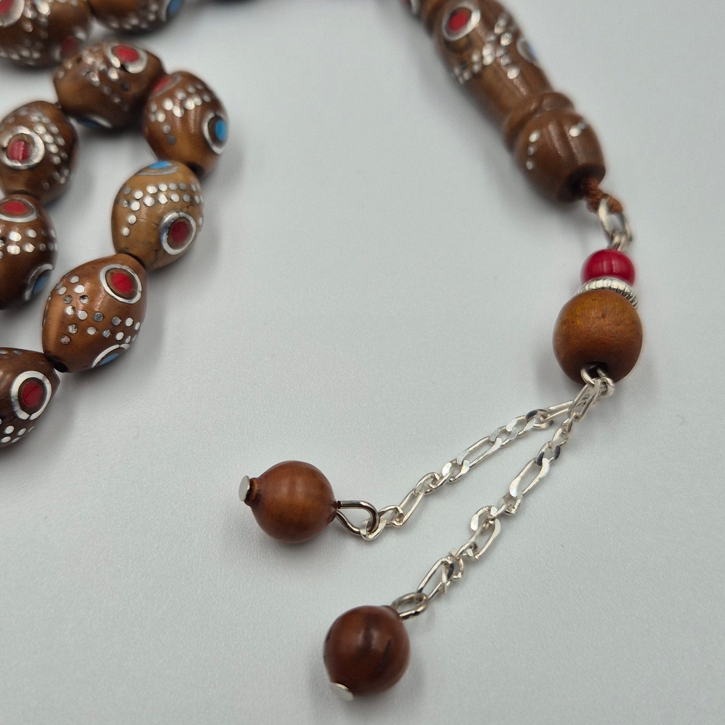 Carved from the resilient Narjil (Coconut) Shell, this tasbih combines organic warmth with metallic precision. Each bead is inlaid with striking Silver-Tone with Red and Blue Craftings.
