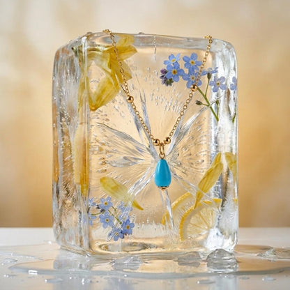 Fairouz teardrop pendant displayed in a clear block with pressed blue flowers — December birthstone necklace gift